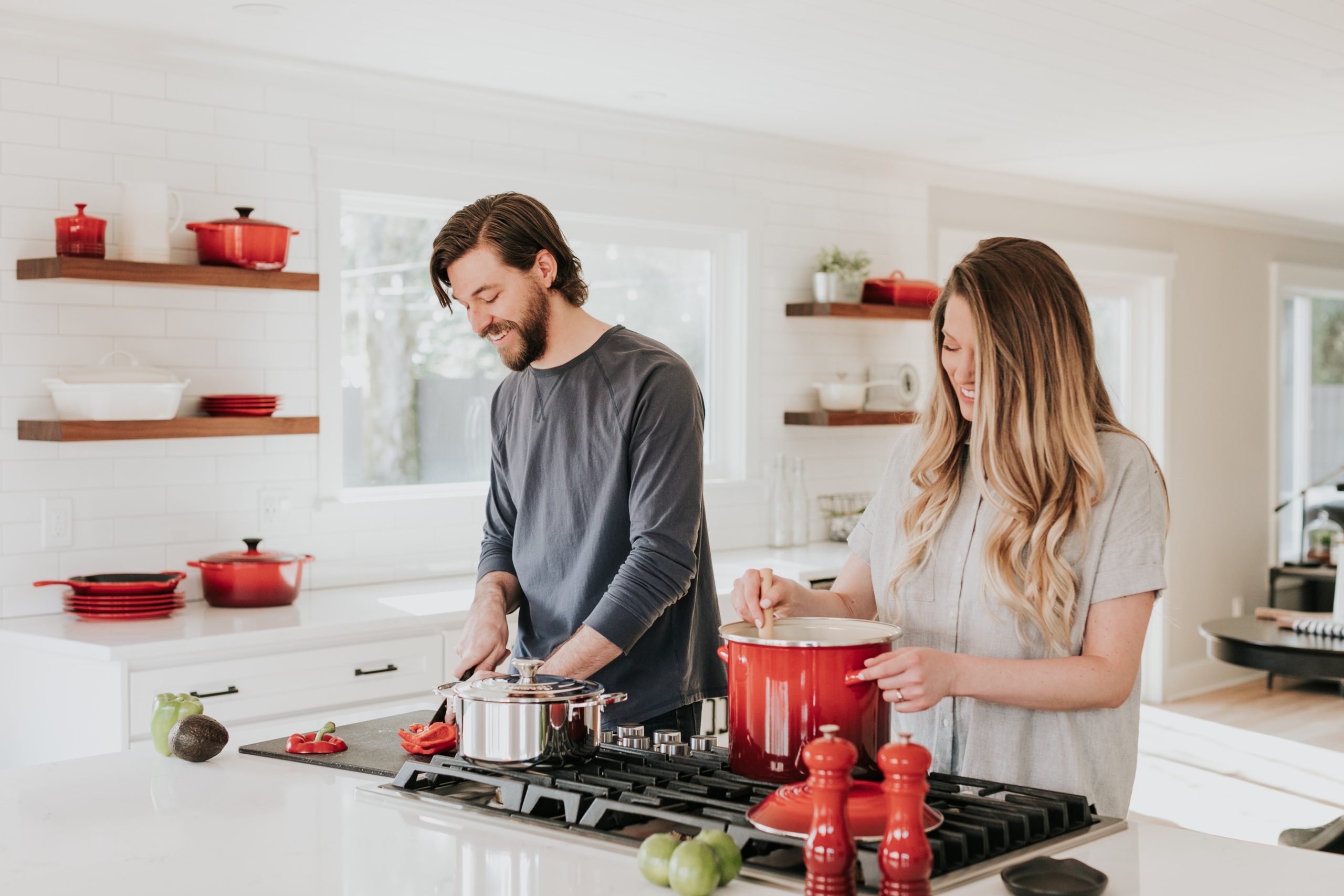 A man smiles as he chops a tomato on the kitchen counter next to his wife who smiles as she's stirring a pot on the stove.