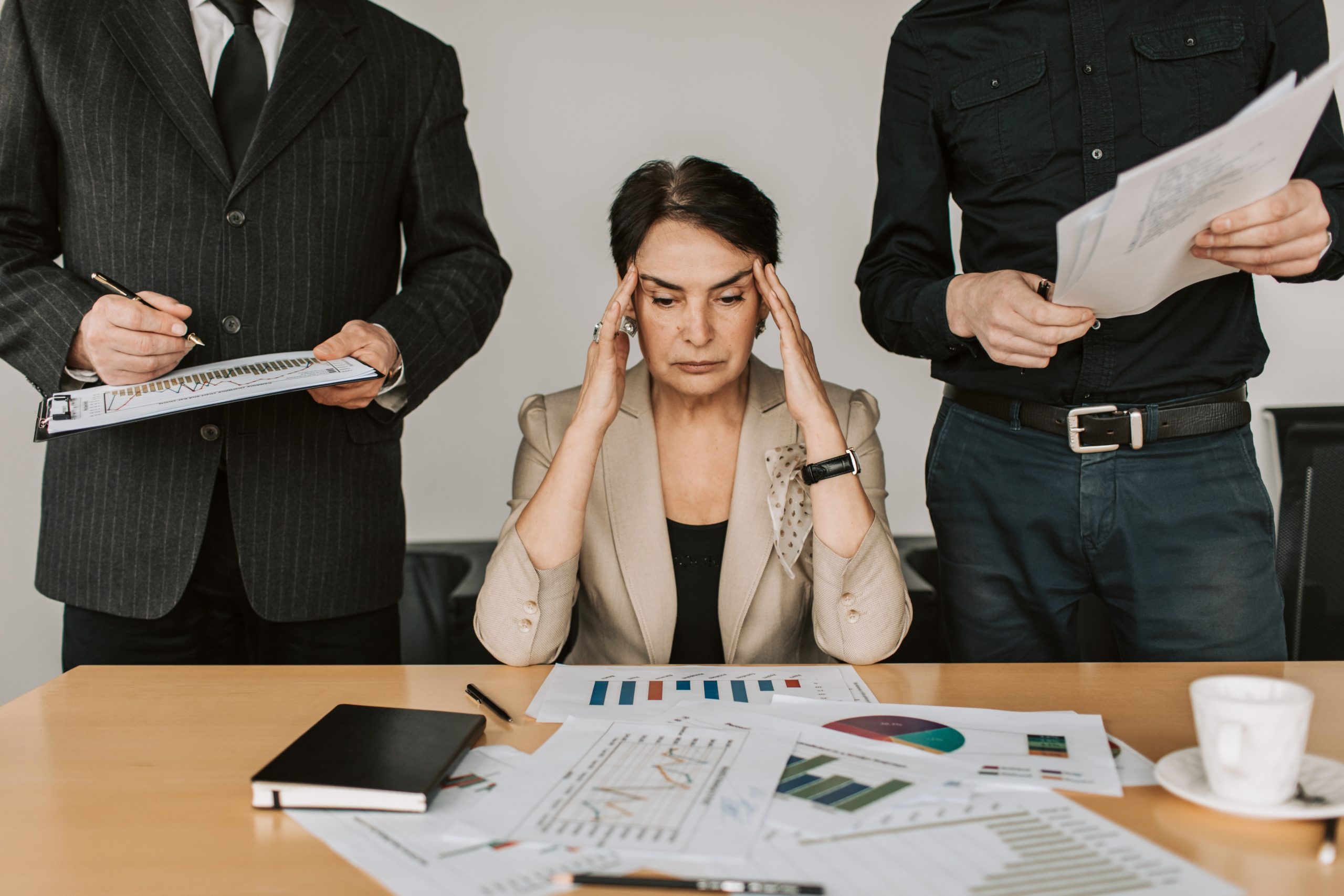 Overwhelmed woman holds her head in her hands, looking down at a mess of papers as men stand on either side of her with papers in their hands as if asking for things